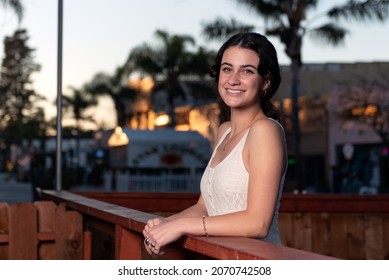 Confident High School Senior Girl Leans On Wooden Handrail In Downtown Ventura With The Golden Sunlight Shiniing Behind Her.