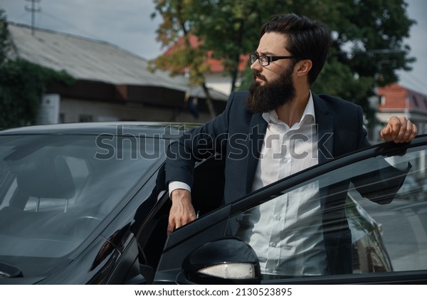 Confident in his choice. Confident man in formal\
wear holding hand on opened car door and looking to the right while\
fixing his glasses with\
hand.