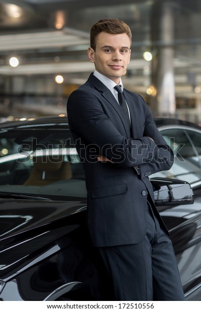 Confident in his choice. Confident man in\
formalwear leaning at the car and looking at camera while standing\
at car dealership