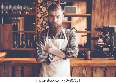 Confident In His New Business. Young Bearded Man In Apron Looking At Camera And Keeping Arms Crossed While Standing At Bar Counter