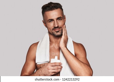 Confident in his skin. Handsome young man applying aftershave lotion and smiling while standing against grey background