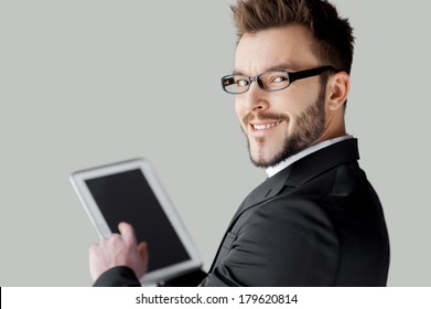 Confident In His Tablet. Rear View Of Cheerful Young Man In Formalwear And Glasses Working On Digital Tablet And Looking Over Shoulder While Standing Against Grey Background