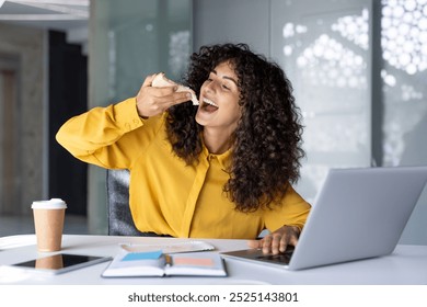 Confident Hispanic businesswoman takes snack break while working at desk with laptop and coffee. Shows balance between work and personal time in modern office environment. - Powered by Shutterstock