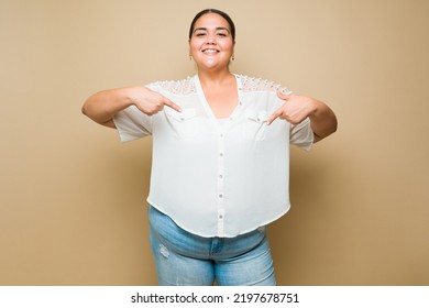 Confident hispanic plus size woman smiling pointing down to copy space ad against a pastel yellow background