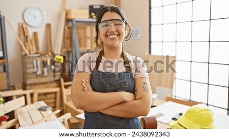Confident hispanic woman with crossed arms standing in a carpentry workshop, wearing safety goggles.