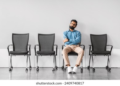Confident indian businessman with a beard, sitting cross-armed in a vacant conference area, exuding a sense of authority and anticipation in a sleek, modern environment
