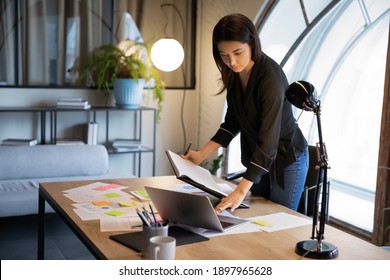Confident Indian businesswoman working with financial project statistics, checking documents with colorful stickers, standing at table in office, female executive using laptop, looking at screen