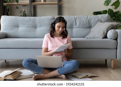 Confident Indian Woman In Headset Translate Document Using Web App Printed Books Making Notes To Paper Copybook. Arab Female Student Sit On Warm Floor At Home Prepare Work Write Up Data From Pc Screen