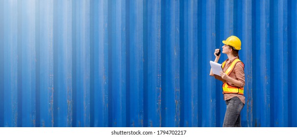 Confident inspector standing in front of cargo container in shipping yard. Smart female engineer, dock worker wear hard hat using clipboard and radio to command transportation. Concept of logistics