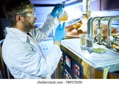 Confident inspector wearing white coat and protective goggles holding flask with beer in hands while checking its purity - Powered by Shutterstock