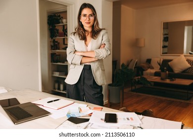 Confident Interior Designer Standing In Her Home Office With Her Arms Crossed. Young Businesswoman Looking At The Camera While Standing Behind Her Desk. Female Designer Planning A New Project Remotely