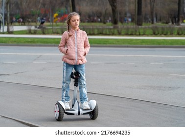 Confident Kid Girl Stand And Ride Near Road On Modern Eco-friendly Electric Self-balancing Scooter. Electric Hoverboard On City Street.