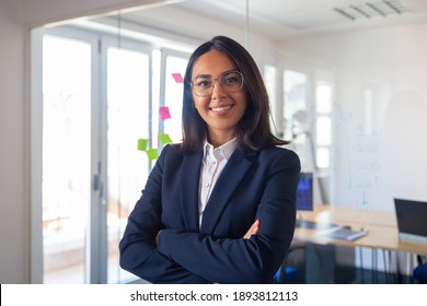 Confident Latin Business Leader Portrait. Young Businesswoman In Suit And Glasses Posing With Arms Folded, Looking At Camera And Smiling. Female Leadership Concept