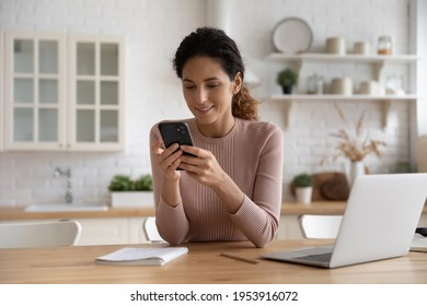 Confident latin female remote employee freelancer sit at kitchen table search files on phone to forward on laptop via home wireless network. Smart young lady user sync restore data using two gadgets