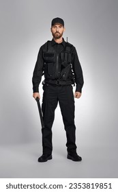 Confident law enforcement officer in black uniform standing with baton in studio. Front view of bearded policeman holding truncheon and looking at camera, on gray background. Police equipment concept.