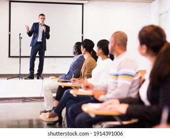 Confident lecturer standing with microphone on stage in conference room, speaking to businesspeople at seminar