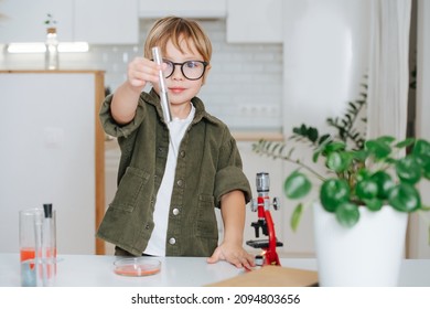 Confident Little Boy Doing Science Project, Showing A Flask With Clear Liquid, Holding It In Outstretched Hand. Behind A Table.