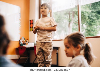 Confident little boy stands in front of his class to give a speech in a language class. Male elementary school pupil develops his public speaking skills during an oral lesson. - Powered by Shutterstock