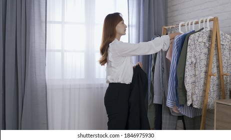 Confident Looking Asian Woman Getting Dressed By Clothing Rack, Swiftly And Elegantly.  Female Ceo Slipping On Black Suit Jacket And Straightening The Collar And Sleeves. Authentic Lifestyle