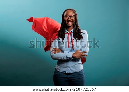 Confident looking mighty powerful young adult person wearing superhero costume on blue background. Portrait of happy brave woman with superpowers wearing red hero cape while standing with arms