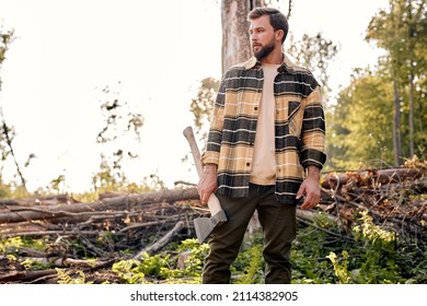 Confident lumberjack works in summer forest. Lumberjack with an ax in the evening. Wood man holding an ax. Young caucasian woodcutter in plaid shirt posing, looking at side in contemplation