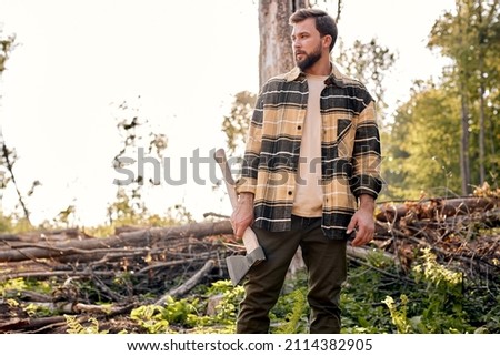 Confident lumberjack works in summer forest. Lumberjack with an ax in the evening. Wood man holding an ax. Young caucasian woodcutter in plaid shirt posing, looking at side in contemplation