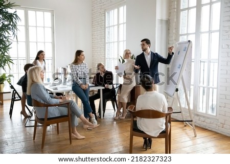 Confident male boss or trainer make whiteboard presentation for diverse employee at team office meeting. Businessman talk lead briefing, present project on flip chart at team training or briefing.