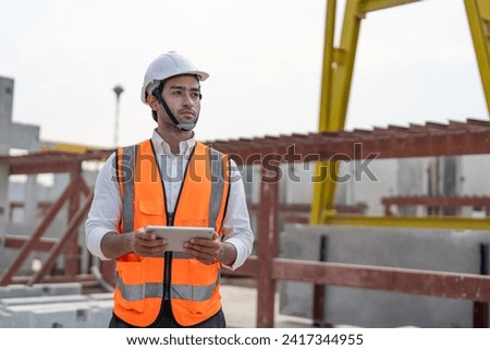 Confident male civil engineer wearing safety vest with white helmet standing and holding tablet working at factory or construction site making precast concrete wall for real estate housing