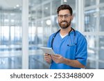 Confident male doctor in blue scrubs holding tablet in modern hospital setting. Professional healthcare worker with stethoscope, embracing technology for patient care and communication.