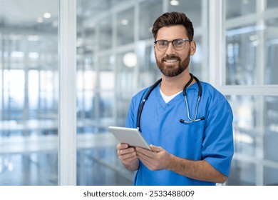 Confident male doctor in blue scrubs holding tablet in modern hospital setting. Professional healthcare worker with stethoscope, embracing technology for patient care and communication. - Powered by Shutterstock