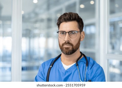 Confident male doctor wearing scrubs and glasses stands in hospital corridor. He has stethoscope around neck, expressing professionalism and readiness. Modern medical environment surrounds. - Powered by Shutterstock