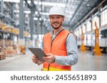 A confident male engineer stands with arms crossed in a large train maintenance hall, wearing a safety helmet and vest.