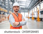 A confident male engineer stands with arms crossed in a large train maintenance hall, wearing a safety helmet and vest.