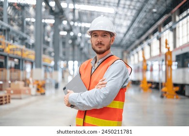 A confident male engineer stands with arms crossed in a large train maintenance hall, wearing a safety helmet and vest. - Powered by Shutterstock