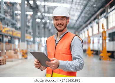 A confident male engineer stands with arms crossed in a large train maintenance hall, wearing a safety helmet and vest. - Powered by Shutterstock