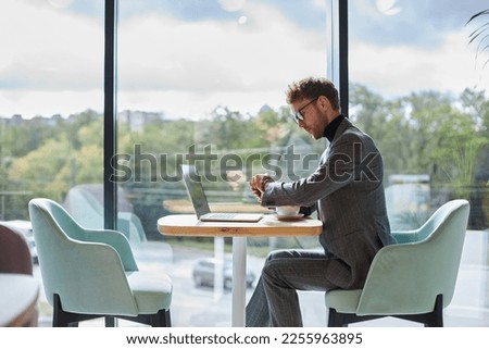 Confident male entrepreneur in formal suit, sitting at a desk in stylish modern light office, working on laptop, checking the time on his wristwatch in anticipation of a meeting with business partners
