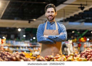 Confident male grocery store worker wearing apron standing with arms crossed in produce aisle. Fresh fruits and vegetables in foreground. Bright store lighting creates warm, inviting atmosphere - Powered by Shutterstock