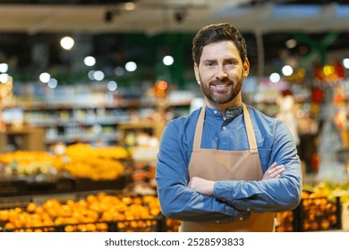 Confident male grocery worker wearing apron stands with crossed arms in front of fresh produce section at supermarket. Smiling expression conveys professionalism, hospitality, dedication. - Powered by Shutterstock