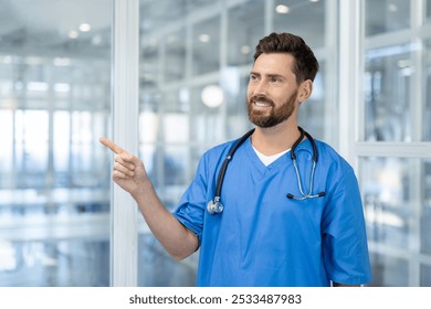 Confident male healthcare professional in blue scrubs with stethoscope indoors. Smiling, pointing doctor embodies medical expertise, positive energy, teamwork in healthcare. Bright, modern setting. - Powered by Shutterstock