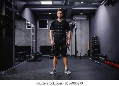 A confident male instructor dressed in a special suit for EMS technology stands in the gym with a dark gray atmosphere. Revolution in training, body rehabilitation, fitness workers and staff