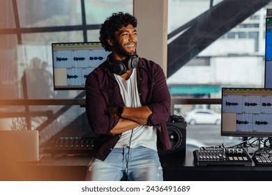 Confident male music producer stands with arms crossed in a modern sound recording studio, with mixers, monitors, and audio workstations.  - Powered by Shutterstock