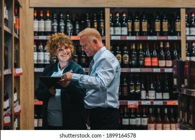 Confident Male Sommelier And Colleague Discussing Various Sorts Of Wine In Cellar. Liquor Store Worker With Female Employee Taking Inventory.