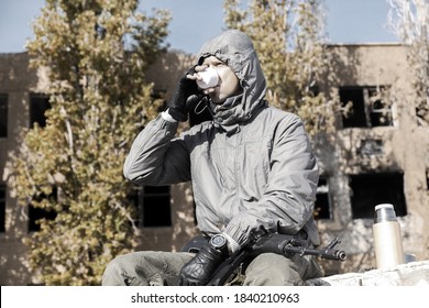 A Confident Male Stalker In A Jacket With A Hood Sits And Drinks Thermos Tea, Weapons On His Lap, Against The Background Of A Ruined House Outside