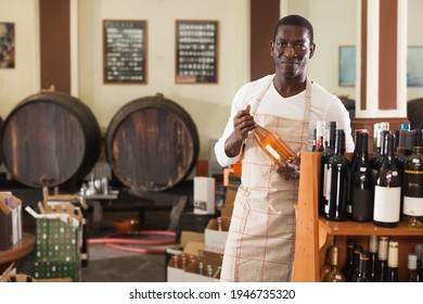 Confident Male Vintner Choosing A Wine Bottle On Shelf Rack And Proffering To Buy At Modern Wine Shop