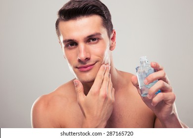 Confident Man Applying Lotion After Shave On Face Over Gray Background. Looking At Camera