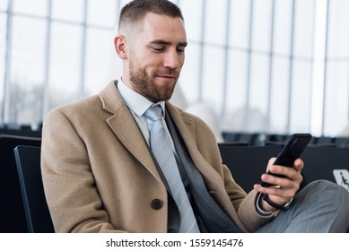 Confident Man Boss In Luxury Suit Is Checking E-mail Via Cellphone Before Meeting With Staff. Male Professional Employer Is Typing Text Message On Mobile Phone, While Is Sitting In Airport Hallway