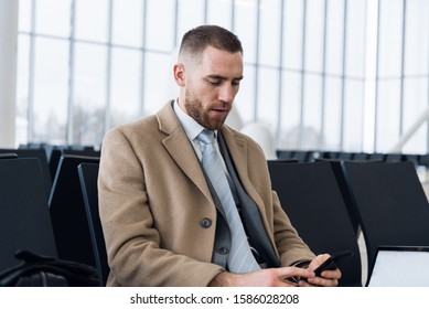 Confident Man Boss In Luxury Suit Is Checking E-mail Via Cellphone Before Meeting With Staff. Male Professional Employer Is Typing Text Message On Mobile Phone, While Is Sitting In Airport Hallway