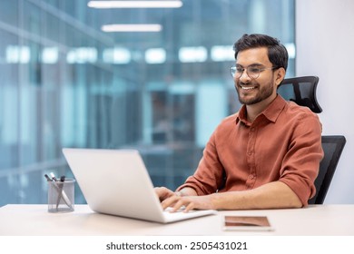 Confident man in casual attire working on laptop at office desk, with phone nearby. Bright, modern workspace reflects productivity and comfort. - Powered by Shutterstock