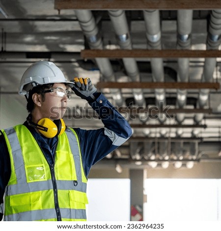 Confident man construction worker with reflective vest, protective goggles and ear muffs. Asian male maintenance engineer holding his safety helmet while looking at piping system at construction site