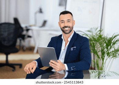 A confident man dressed in a stylish striped suit sits at a desk in a contemporary office. He smiles while holding a tablet, surrounded by a clean, organized workspace. - Powered by Shutterstock